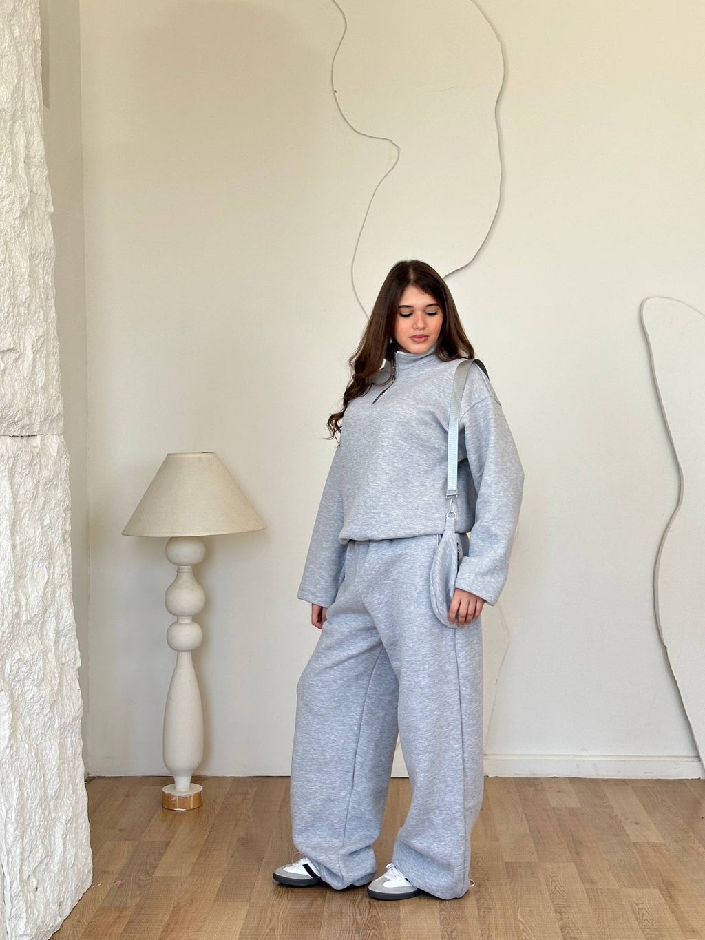 Woman wearing a gray outfit standing in a room with a lamp and decorative wall.